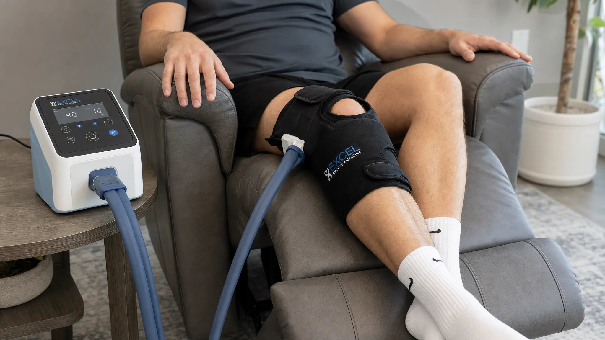 A cold compression device connected by tubing to an anatomical knee wrap on a reclined patient, with the control unit positioned on a small table beside the chair.