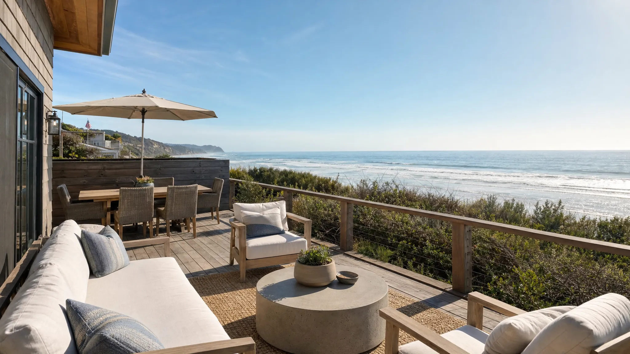 A quiet coastal terrace with lounge seating, a small dining table, and the ocean visible beyond dunes or a bluff, showing a private beach rental's outdoor space.