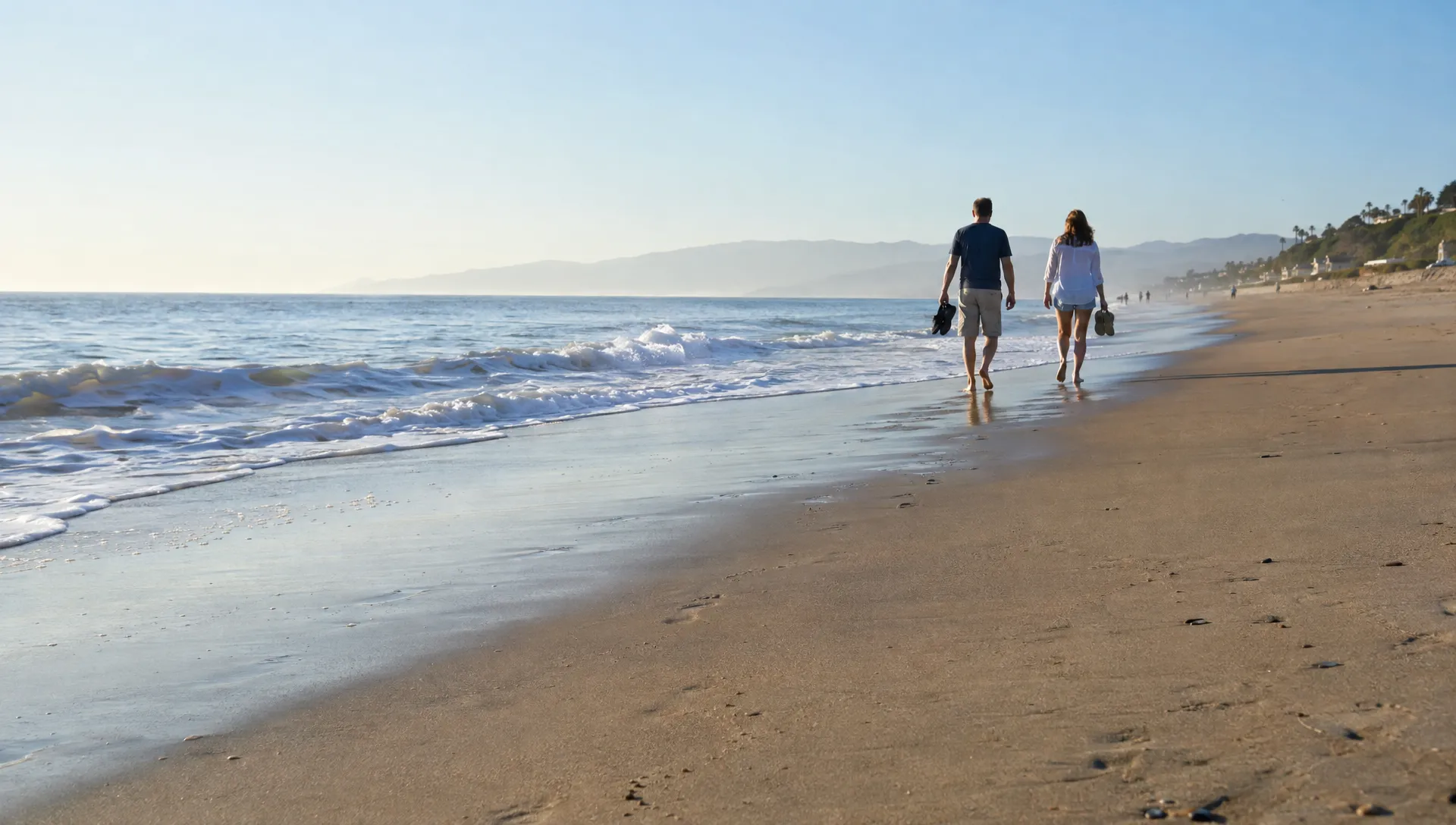 Two companions carry their shoes along the water's edge on a quiet morning beach walk.