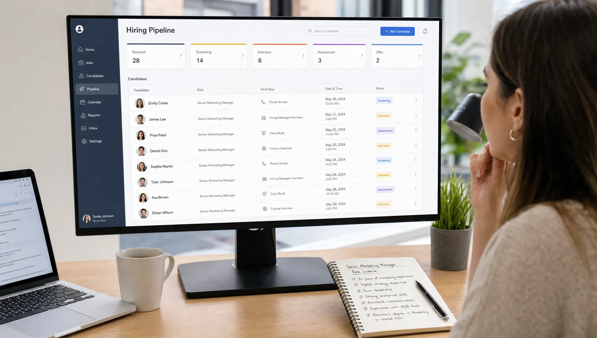 A recruiter reviews an organized hiring pipeline on a dashboard with candidate profiles, interview times, and status updates, while a notebook with role criteria sits beside the workstation.