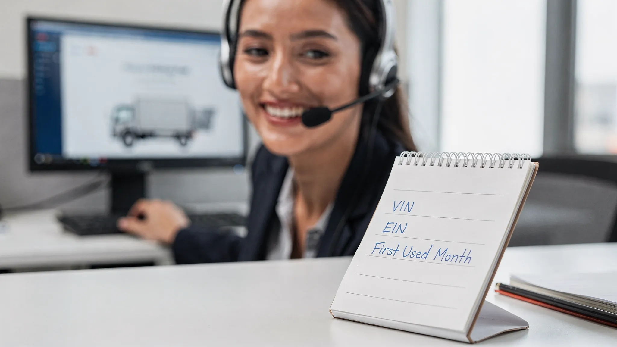 A bilingual customer support specialist wearing a headset, smiling while assisting a truck driver over the phone, with a notepad showing VIN, EIN, and First Used Month details.
