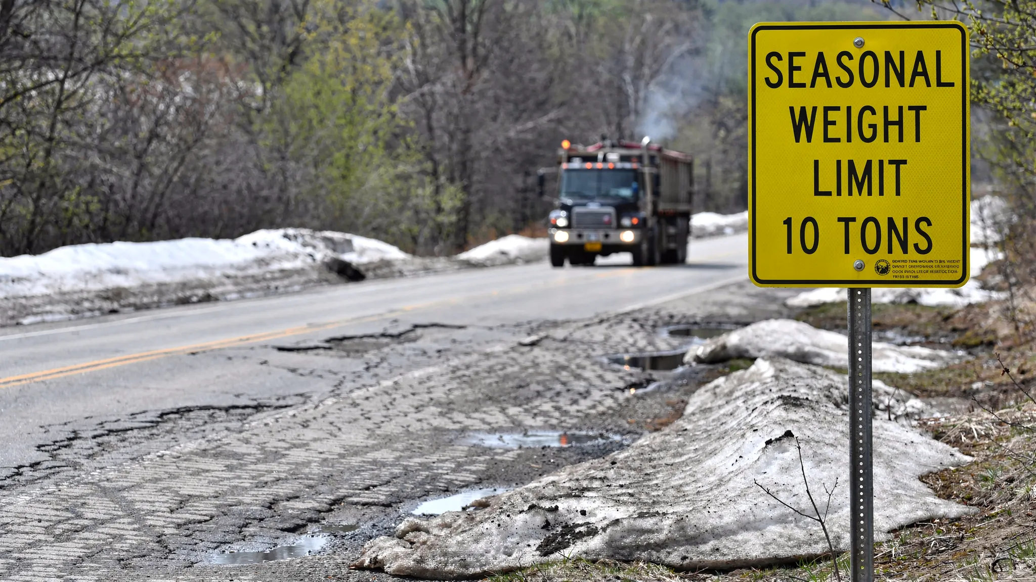 A winter-to-spring road scene showing a posted seasonal weight restriction sign near a frost-heaved roadway surface, with a heavy truck in the distance and subtle pavement cracking visible.